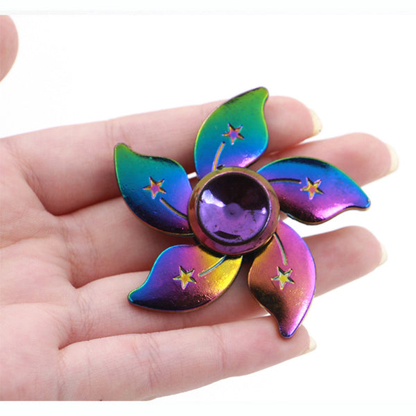 Toupie anti stress en métal tenu dans une main, la toupie est en forme de fleur et est de couleur arc-en-ciel.